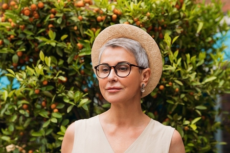 Stylish mature woman with grey hair wearing a straw hat and looking down while standing outdoors