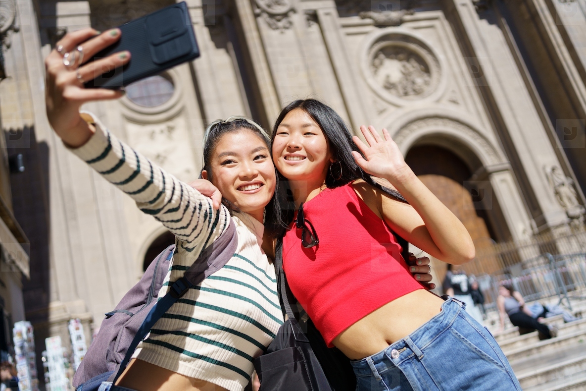 Happy Asian female tourists taking self portrait on smartphone against Cathedral of Granada in Spain