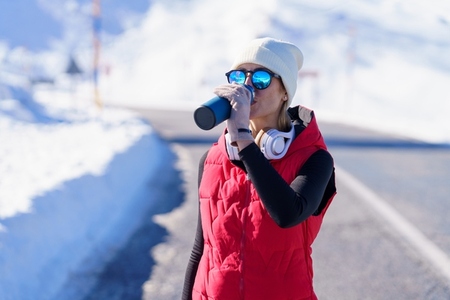 Athletic woman in outerwear drinking water in snowy terrain