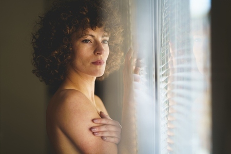 Pensive young nude woman looking away  leaning on window
