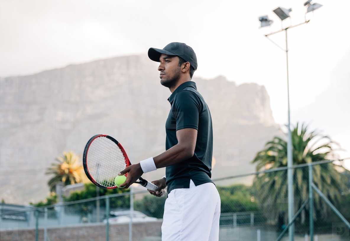 Fit man in sportswear and cap with a racket standing on tennis court  Tennis player looking away while standing on a hard court