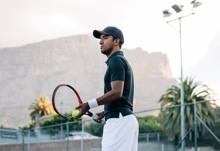 Fit man in sportswear and cap with a racket standing on tennis court  Tennis player looking away while standing on a hard court