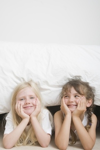 Two young girls with chin resting on hands under bed smiling