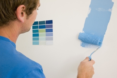 Back view of man holding paint roller and painting a white wall with blue paint