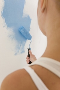Back view of woman holding paint brush and painting a white wall with blue paint