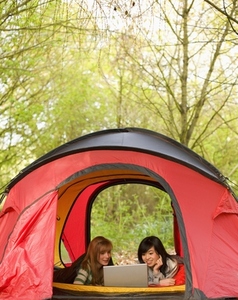 Two smiling teenaged girls lying in a tent looking at laptop computer screen