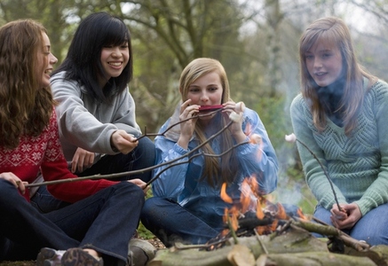 Teenage girls roasting marshmallow over campfire one is playing the harmonica