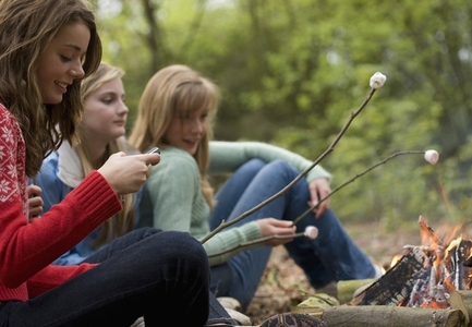 Teenage girls roasting marshmallow over campfire one is using a cell phone