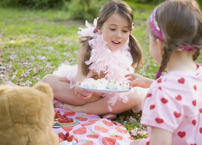 Young girl sitting in a garden lawn wearing a pink feather boa offering cupcakes to a friend