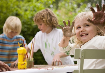 Young girl sitting at table holding arms up with hands covered in paint