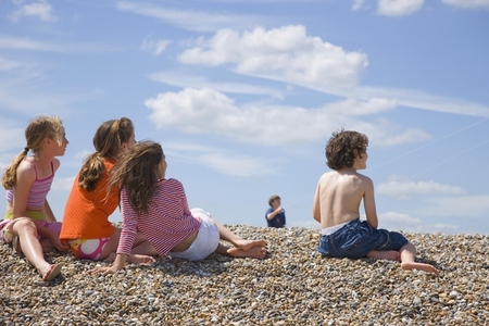 Children sitting on a beach watching a  young boy flying a kite