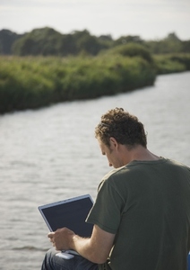 Back view of a man sitting by a river using a laptop computer