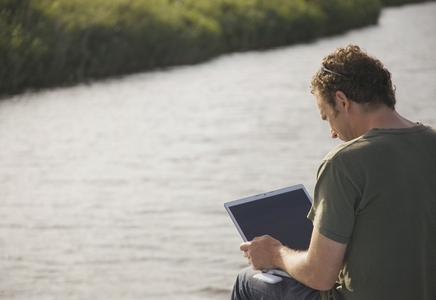 Back view of a man sitting by a river using a laptop computer
