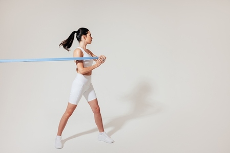 Slim female in white fitness attire pulling resistance band at the white wall in a studio
