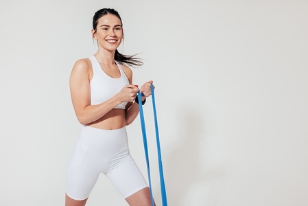 Cheerful female in white fitness attire exercising with blue resistance band at a white wall