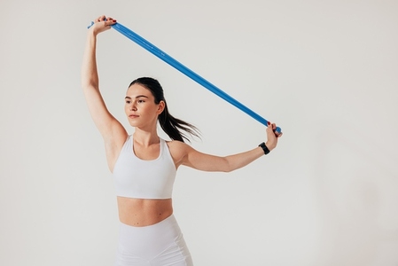 Young fit woman in white sportswear exercising with resistance band at white wall
