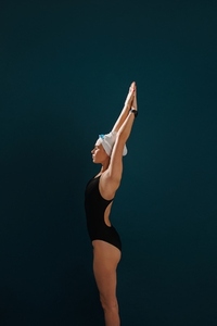 Side view of a young slim female swimmer preparing for competition standing against a blue backdrop