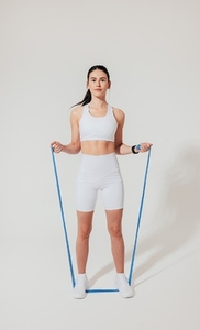 Full length of a young female in white fitness clothes practicing with a resistance band on a white backdrop