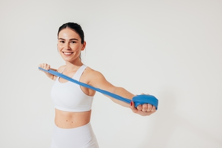Cheerful woman in white fitness wear is warming up her arms with a resistance band in the studio