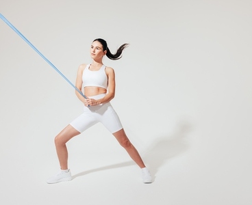 Young slim woman in white fitness wear practicing on a white backdrop with a resistance band
