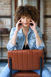 Woman in denim jacket and bra sitting on chair and touching face