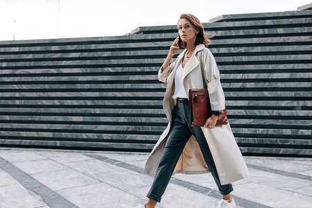 Stylish woman in a coat walking outdoors talking on a mobile phone  Businesswoman with ginger hair and a leather folder walking on the street