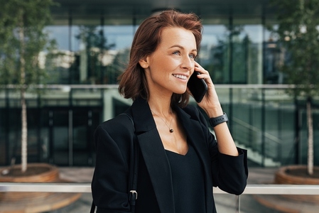 Smiling middle aged woman talking on mobile phone at business building  Cheerful female with ginger hair in formal clothes standing outdoors and talking on her cell phone