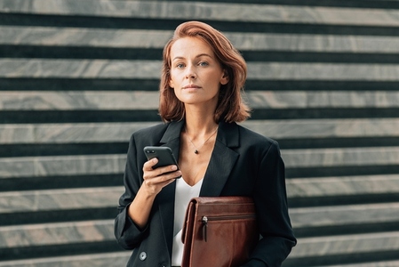 Middle aged businesswoman holding a leather folder and smartphone and looking at camera while standing outdoors