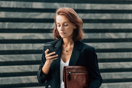 Confident middle aged female with a leather folder looking at her smartphone while standing outdoors