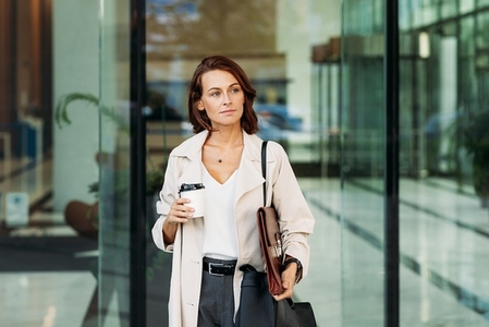 Middle aged businesswoman holding coffee going out from the business building  Confident businesswoman with ginger hair at business building