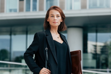 Confident middle aged business woman with bag and leather folder standing against office building