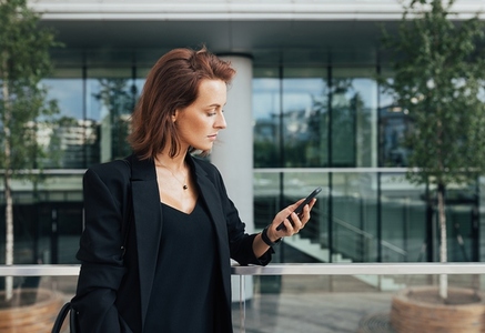 Side view of a confident businesswoman with ginger hair looking at her smartphone against an office building