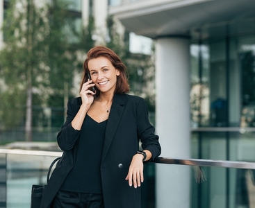 Cheerful businesswoman with ginger hair talking on a mobile phone  Smiling middle aged female in formal clothes making a phone call outdoors