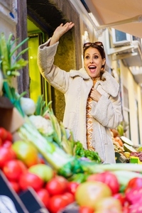 Excited female greengrocer in cozy coat waving hand in fruits shop