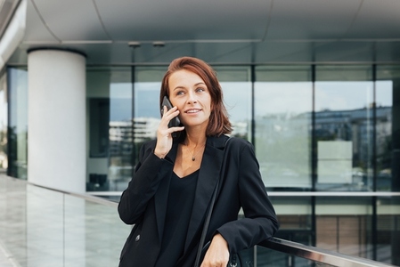 Smiling middle aged business woman with ginger hair leaning on a railing talking on a mobile phone