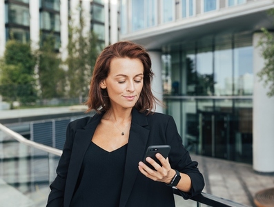 Middle aged businesswoman looking at her mobile phone while leaning a railing against an office building