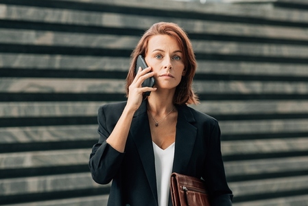 Businesswoman with ginger hair in formal wear making a phone call while standing outdoors