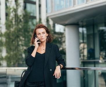 Confident middle aged businesswoman with ginger hair leaning on railing talking on mobile phone outdoors