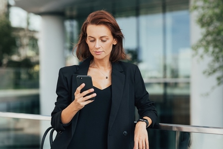Middle aged businesswoman in black formal wear looking at her smartphone while standing outdoors