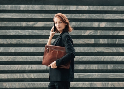 Side view of a confident middle aged female with a leather folder  Businesswoman with ginger hair making a phone call while standing outdoors