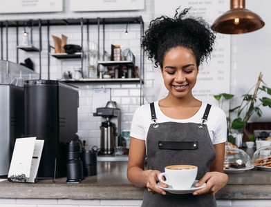 Smiling barista in an apron looking at a cup of coffee  Woman with curly hair works as a barista in a coffee shop