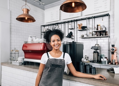 Young smiling woman with curly hair working as a barista  Female in an apron standing at a counter in a coffee shop and looking at camera