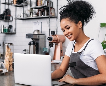 Smiling woman with curly hair wearing an apron typing on a laptop in a coffee shop