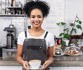 Smiling waitress with cup of coffee  African American woman working as a barista holding a cup with a freshly prepared cappuccino