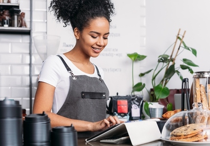 Barista in apron typing on digital tablet  Cheerful woman with curly hair working as a barista in a coffee shop using a digital tablet