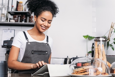 Side view of smiling waitress with curly hair typing on a digital tablet in a coffee shop
