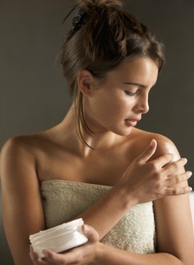 Young woman wrapped in a towel holding cream jar and rubbing her shoulder with body lotion