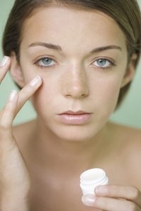 Young woman applying moisturizer under her eye holding a cream  jar