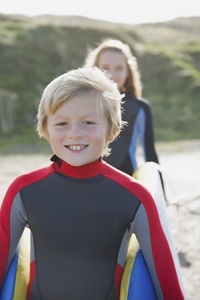 Smiling boy in a wetsuit carrying a surfboard with a girl