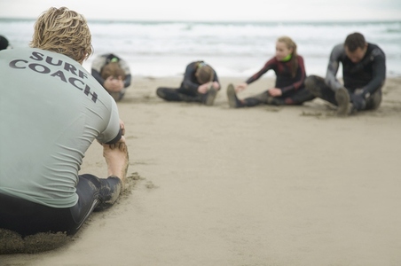 Back view of a surfing instructor and students stretching their legs on a beach
